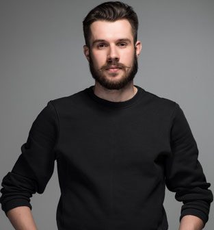 A man with beard and black shirt standing in front of gray background.